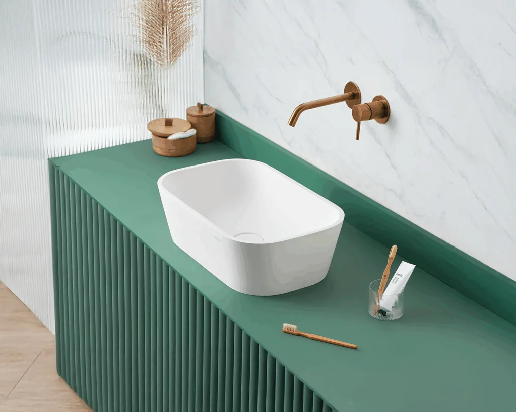 White basin on a green countertop, copper faucet, two wooden boxes, and a cup with a toothbrush and toothpaste.