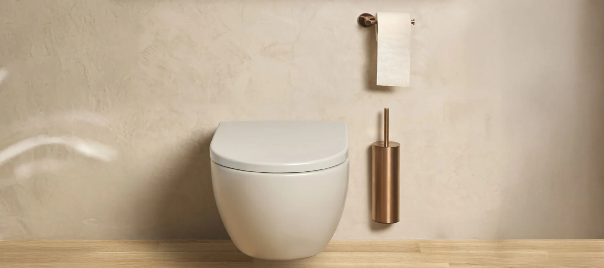 Wall-mounted modern white toilet with brushed copper toilet brush holder and matching toilet paper holder in a minimalist beige bathroom with wooden floor.