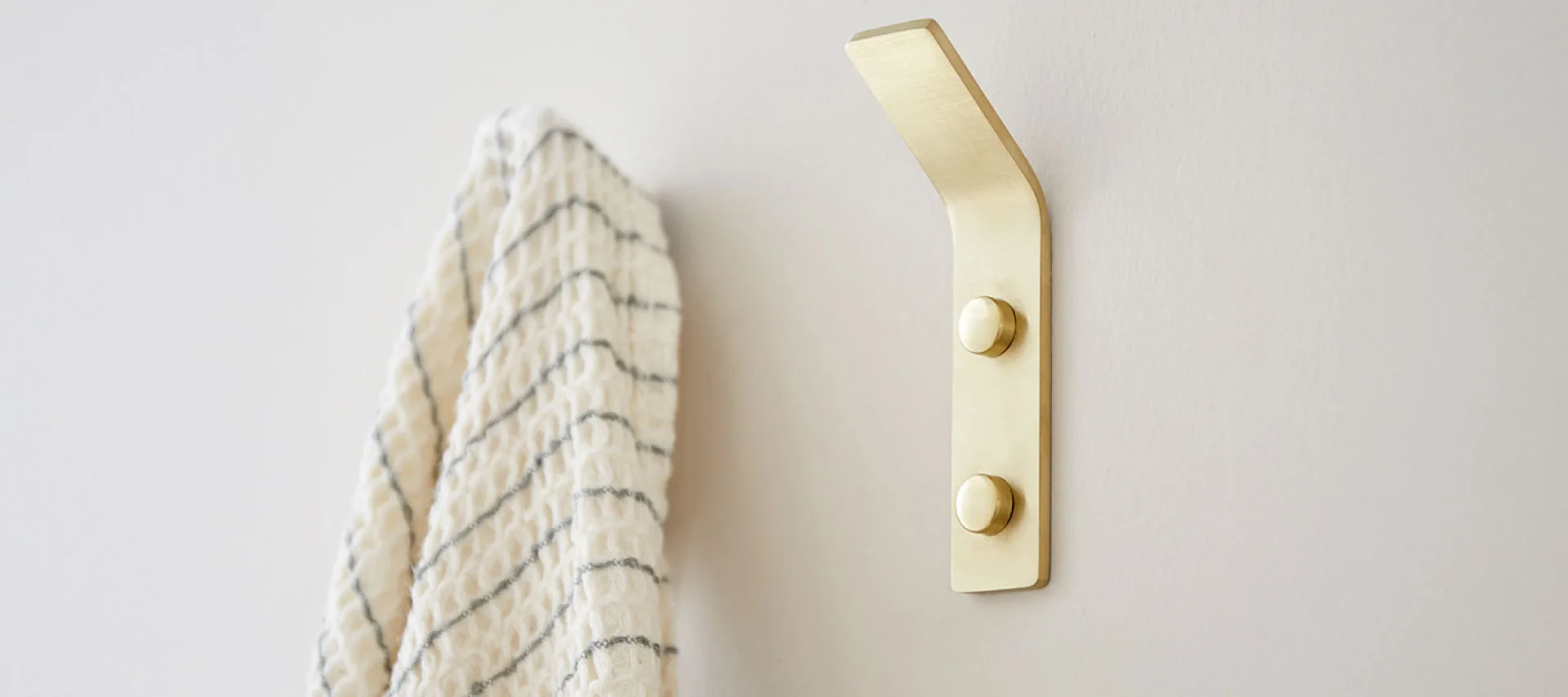Brushed brass wall hook mounted on a white wall holding a cream textured towel with subtle gray stripes in a minimalist bathroom setting.