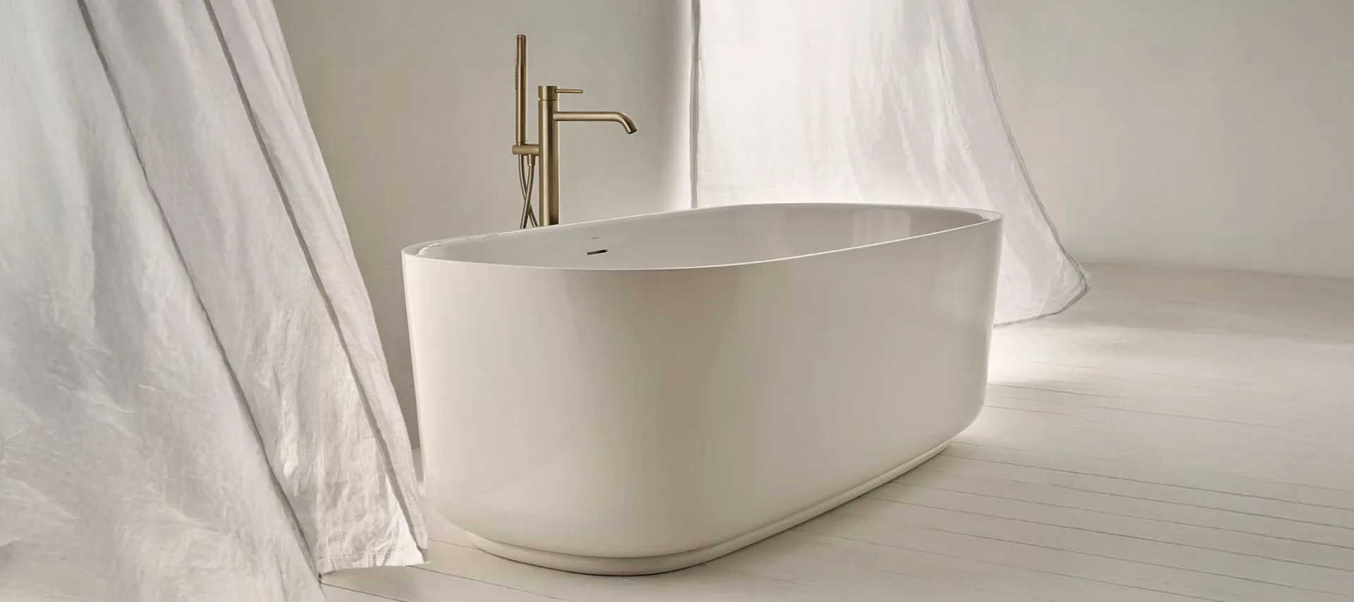 Bathtub mixers : Luxury freestanding white bathtub with brushed gold floor-mounted faucet in a bright minimalist bathroom, featuring soft natural light, sheer white curtains, and light wood flooring for a serene spa-style interior.