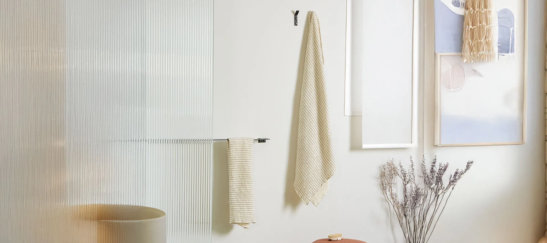 Minimalist spa-inspired bathroom featuring a textured glass shower partition, neutral beige textiles, a terracotta storage stool, dried botanical accents, and soft natural lighting.