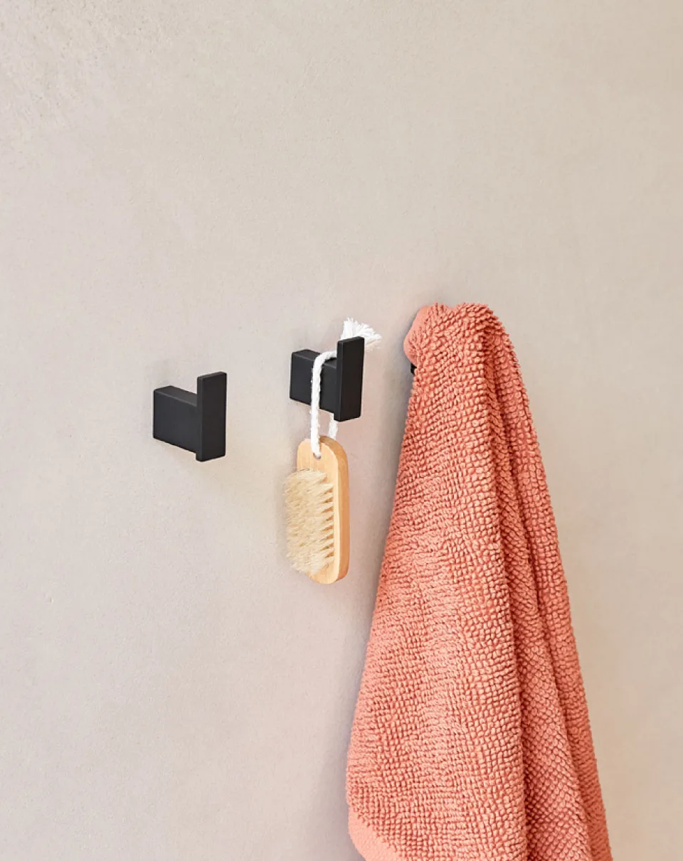 Modern matte grey wall hooks with coral bathroom towel Close-up of matte black wall hooks holding a coral textured towel and a natural wood bath brush against a minimalist beige bathroom wall.